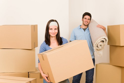 A young woman and a young man, both with light skin and casually dressed in blue shirts, are inside a residential space preparing for a house move. The woman has dark brown hair and is smiling, holding a large, plain cardboard box in front of her with both hands. The man, standing behind her, has short dark hair and is smiling as well, holding a rolled-up beige carpet with a textured surface over his shoulder. The background features a plain white wall, and the scene is filled with several stacked cardboard boxes of varying sizes, positioned on the floor and against the wall, indicating an ongoing packing process typical of house removals in the Highbury area. The lighting appears natural and bright, emphasizing the clean, organized atmosphere suitable for professional removals services. The company's name, Highbury Man with Van, is associated with the scene's context, highlighting local expertise in removals for the London N5 postcode area.
