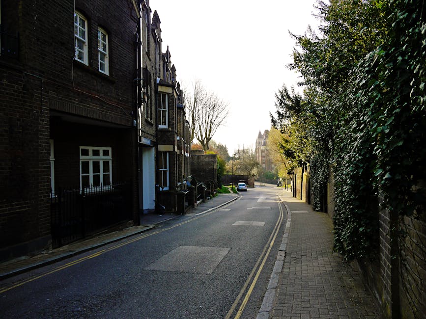 A narrow residential street with multi-storey brick buildings on the left side, featuring white-framed windows and small front gardens behind black metal railings. The street has a gently curved aspha