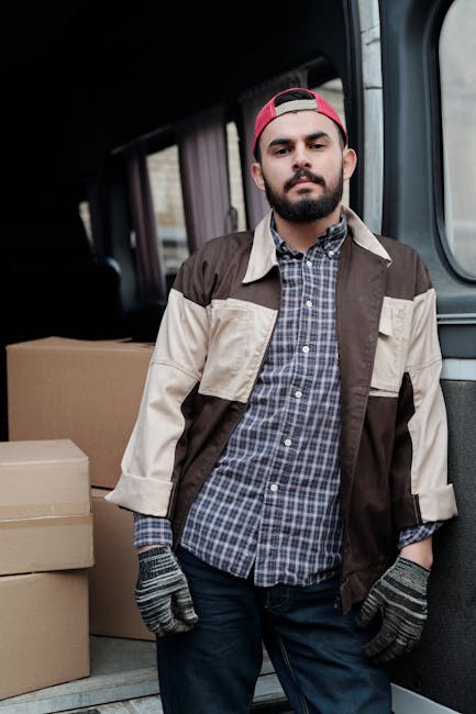 A person wearing a black jacket, dark trousers, and a black cap is inside the open rear doors of a white van loaded with multiple brown cardboard boxes, some with visible labels or stickers, arranged
