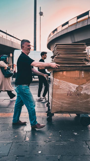 A man is pushing a large metal trolley loaded with multiple flattened cardboard boxes, some wrapped in plastic, on a busy outdoor pavement area under a bridge. He is wearing a dark long-sleeve shirt a